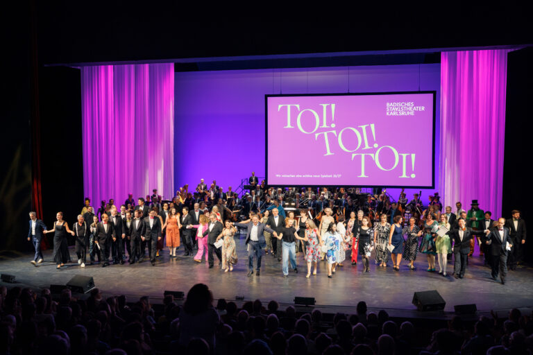 Ensemble und Orchester des Badischen Staatstheaters beim Schlussapplaus, im Hintergrund die Projektion „Toi! Toi! Toi!“